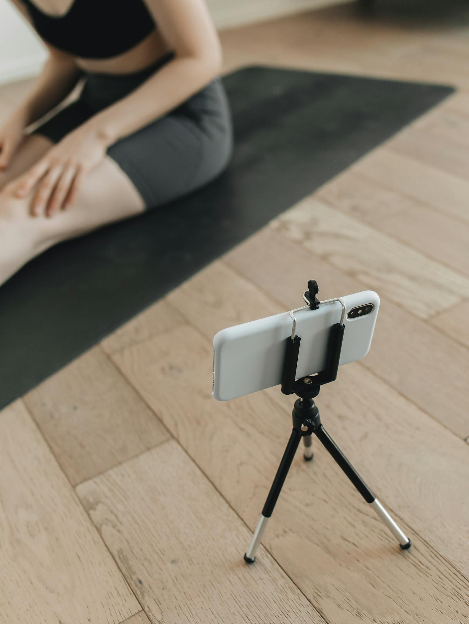 A woman practices yoga indoors, recording herself with a smartphone placed on a tripod.